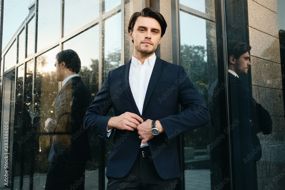 Young handsome bearded brunette man in white shirt and classic jacket standing dreamily looking in camera with glass building on background Young handsome bearded brunette man in white shirt and classic jacket standing dreamily looking in camera with glass building on background