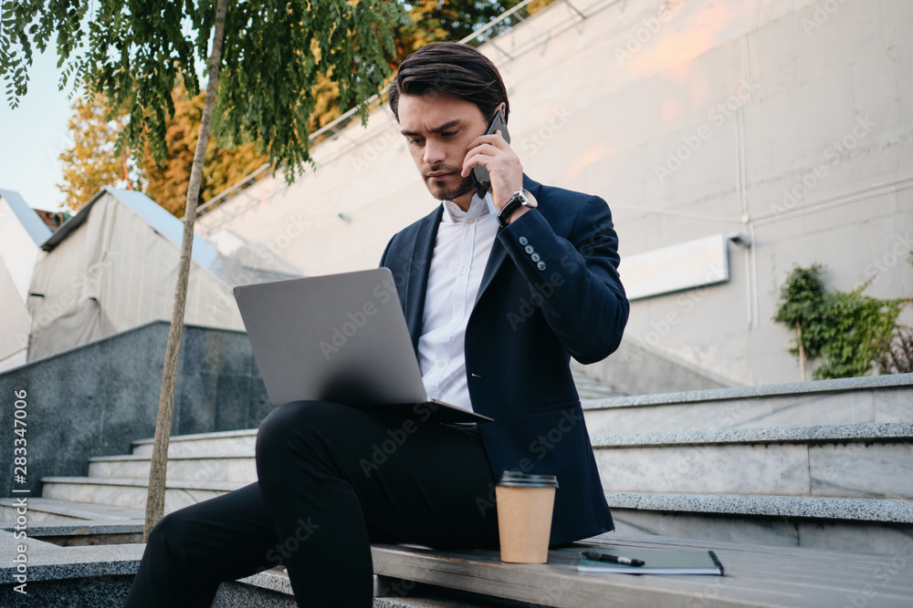 Young handsome bearded brunette man in white shirt and classic suit sitting on bench with cup of coffee to go and laptop on knees thoughtfuly talking on cellphone Young handsome bearded brunette man in white shirt and classic suit sitting on bench with cup of coffee to go and laptop on knees thoughtfuly talking on cellphone