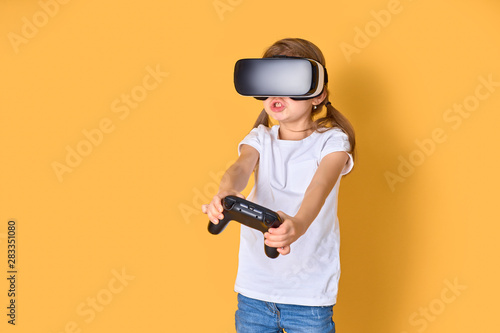 Girl experiencing VR headset vs joystick game on yellow background. Surprised emotions on her face. Child using a gaming gadget for virtual reality. Futuristic goggles at young age. Virtual technology