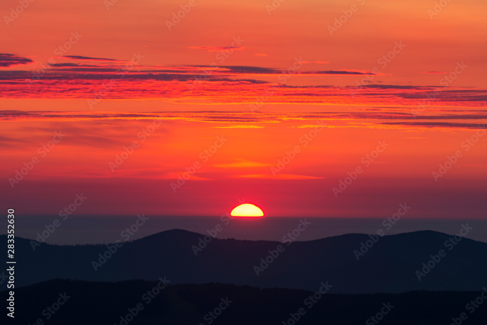 Sunrise in the mountains. The blood red sky is covered with several clouds. Silhouettes of mountains are visible.