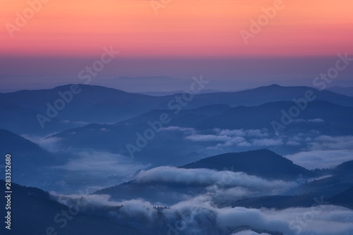 Panoramic view of the ridge of mountain peaks in the early autumn morning. The sky is crimson pink, and clouds swirl in the valley between the mountains. © imartsenyuk