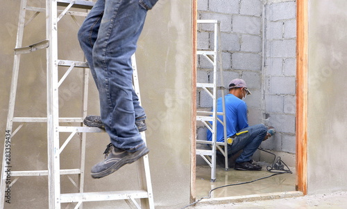 Selective focus at construction worker using angle grinder to working in door frame on cement wall with blurred worker's leg on ladder in foreground 