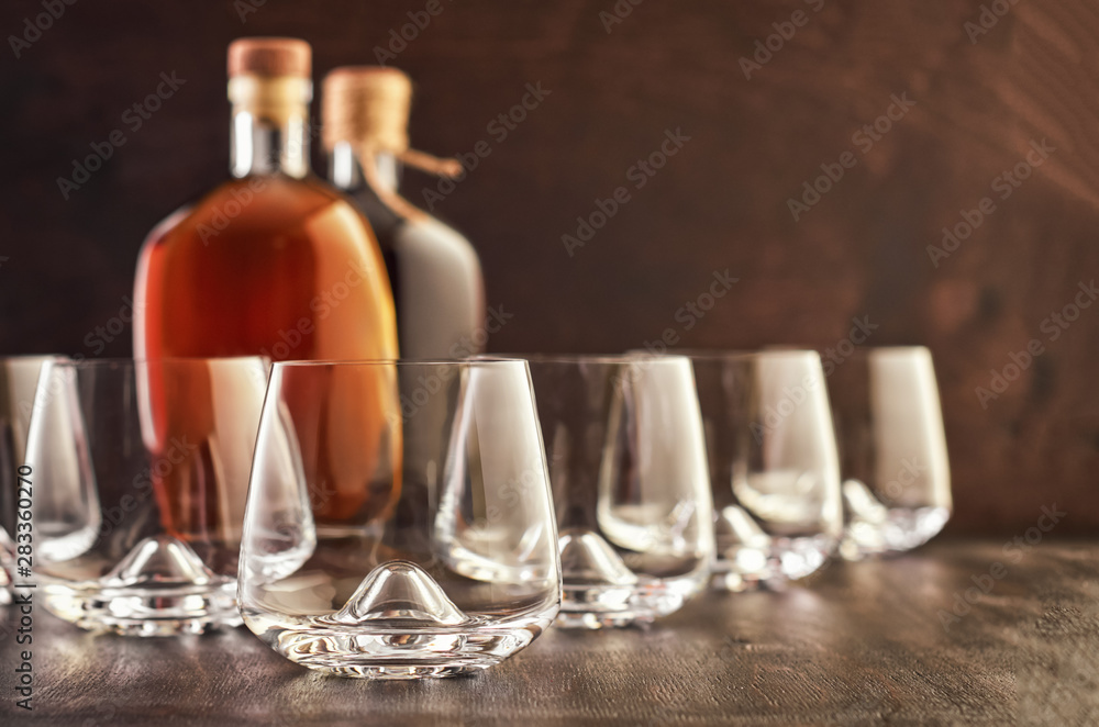 A set of five empty whiskey glasses on a wooden table. In the background are two closed bottles of whiskey. A set of five empty whiskey glasses on a wooden table. In the background are two closed bottles of whiskey.
