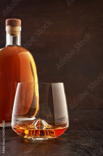A crystal glass of whiskey and a full bottle on a wooden table.
