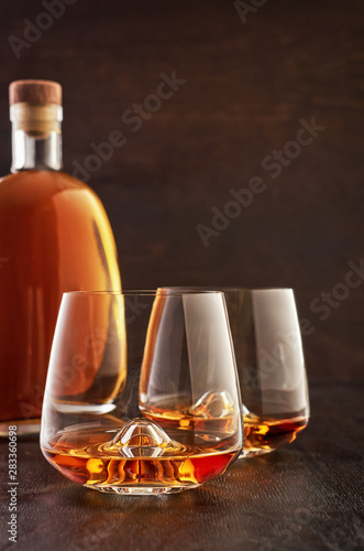 Two crystal glasses of whiskey and a full bottle on a wooden table.