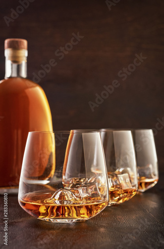 Three crystal glasses of whisky on ice and a full bottle of whisky on a wooden table.