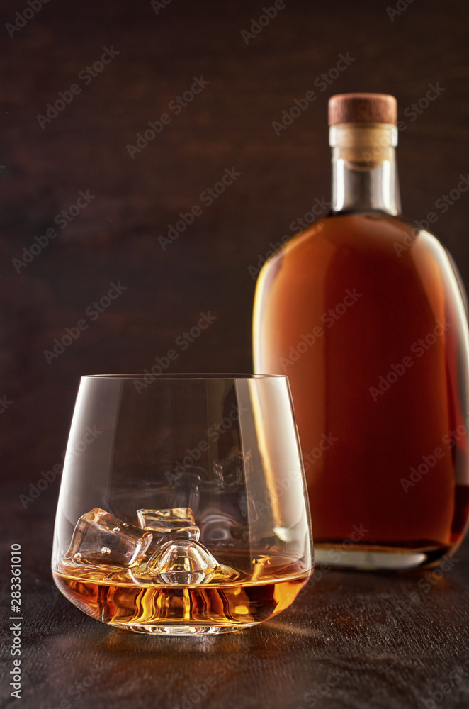 A crystal glass of whisky with ice and a full bottle of whisky on a wooden table. A crystal glass of whisky with ice and a full bottle of whisky on a wooden table.