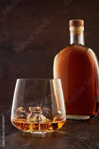 A crystal glass of whisky with ice and a full bottle of whisky on a wooden table.