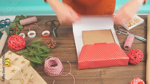 Female hands wrapping Christmas gift in craft ecological paper on dark wooden background with decorating elements, colourfil twine stripes and pack paper rolls. photo