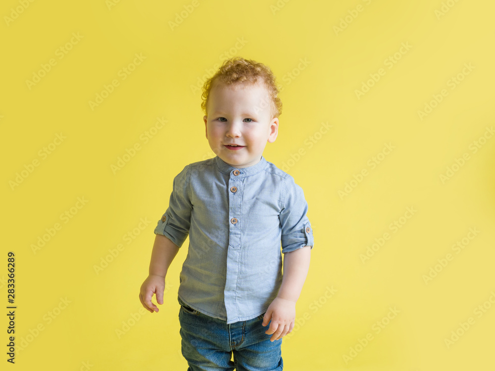 Beautiful red-haired curly baby smiling while standing on a yellow background in a blue shirt and jeans. Studio photography Beautiful red-haired curly baby smiling while standing on a yellow background in a blue shirt and jeans. Studio photography