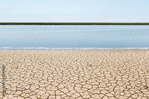 The sunny landscape with straight stripes of the dry cracked soil, fresh surface of water, green opposite bank and blue sky