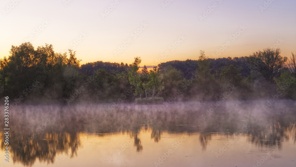 Panorama of tendrils of morning mist on a lake at sunrise and a colorful orange glow in the sky reflected in the tranquil water with the surrounding woodland trees in an atmospheric landscape