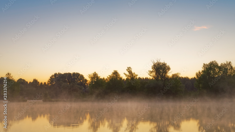 Panorama of tendrils of morning mist on a lake at sunrise and a colorful orange glow in the sky reflected in the tranquil water with the surrounding woodland trees in an atmospheric landscape Panorama of tendrils of morning mist on a lake at sunrise and a colorful orange glow in the sky reflected in the tranquil water with the surrounding woodland trees in an atmospheric landscape
