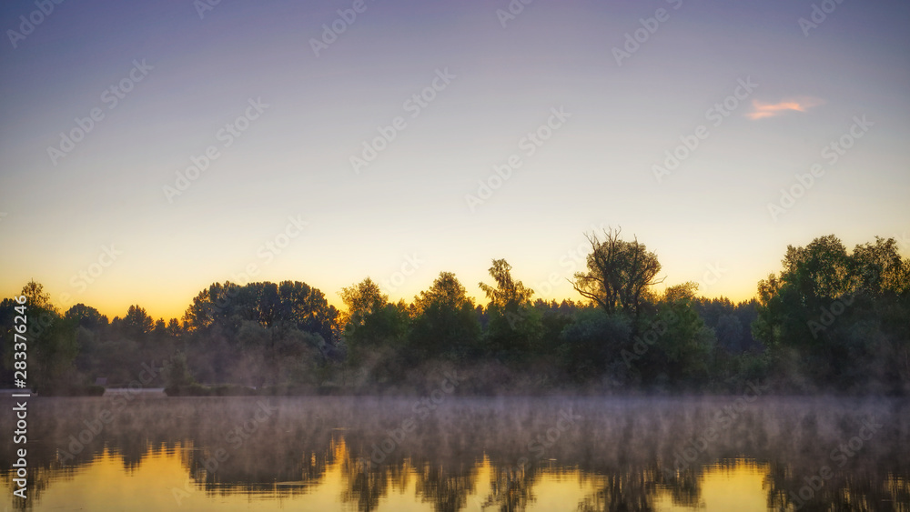 Panorama of tendrils of morning mist on a lake at sunrise and a colorful orange glow in the sky reflected in the tranquil water with the surrounding woodland trees in an atmospheric landscape Panorama of tendrils of morning mist on a lake at sunrise and a colorful orange glow in the sky reflected in the tranquil water with the surrounding woodland trees in an atmospheric landscape