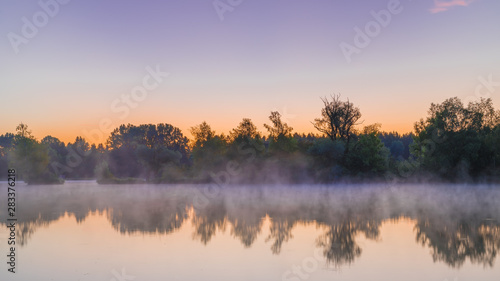 Panorama of tendrils of morning mist on a lake at sunrise and a colorful orange glow in the sky reflected in the tranquil water with the surrounding woodland trees in an atmospheric landscape © Kurt Rabe