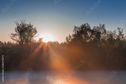 Panorama of tendrils of morning mist on a lake at sunrise and a colorful orange glow in the sky reflected in the tranquil water with the surrounding woodland trees in an atmospheric landscape