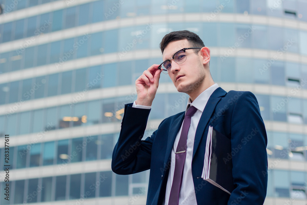 Serious self-confident bold young businessman with a file or folder for documents holding his glasses, standing outdoor office modern building or business centre, looking down at camera in formal suit Serious self-confident bold young businessman with a file or folder for documents holding his glasses, standing outdoor office modern building or business centre, looking down at camera in formal suit