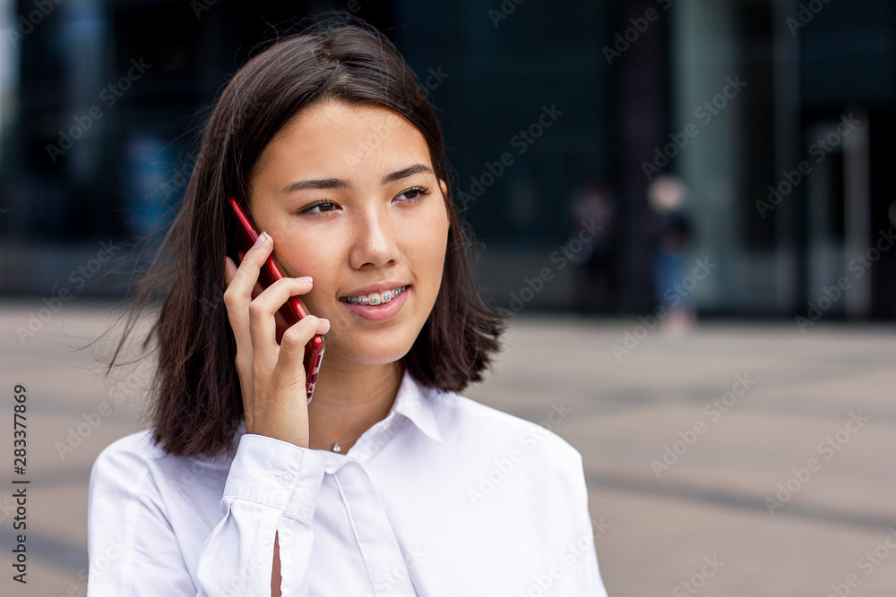 Asian young businesswoman talking on cell phone, formal dressed in white shirt, smiling outside office. Beautiful Chinese girl with braces having business conversation on smartphone outdoor work place Asian young businesswoman talking on cell phone, formal dressed in white shirt, smiling outside office. Beautiful Chinese girl with braces having business conversation on smartphone outdoor work place