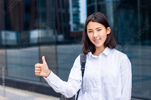 Happy young Asian girl, student, pupil or businesswoman with a backpack smiling,  looking straight at camera and showing thumb up outside. Female standing outdoor office or workplace. Sign of approval © Евгений Шемякин