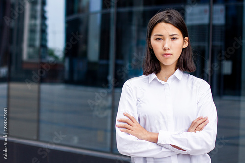 Serious asian young businesswoman or university student, formal dressed in white shirt standing outdoor near office modern building or business centre. Girl with her hands crossed looking at camera.