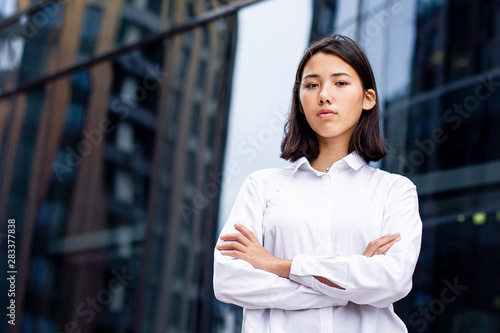 Asian serious young girl standing outdoor business office building with her hands crossed and looking straight at camera in white formal shirt. Chinese or Korean confident female businesswoman
