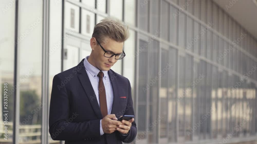 Happy businessman smiling as he reads a text message standing outside the office. Male executive manager in trendy clothes using smartphone outdoors. Modern technology concept Happy businessman smiling as he reads a text message standing outside the office. Male executive manager in trendy clothes using smartphone outdoors. Modern technology concept