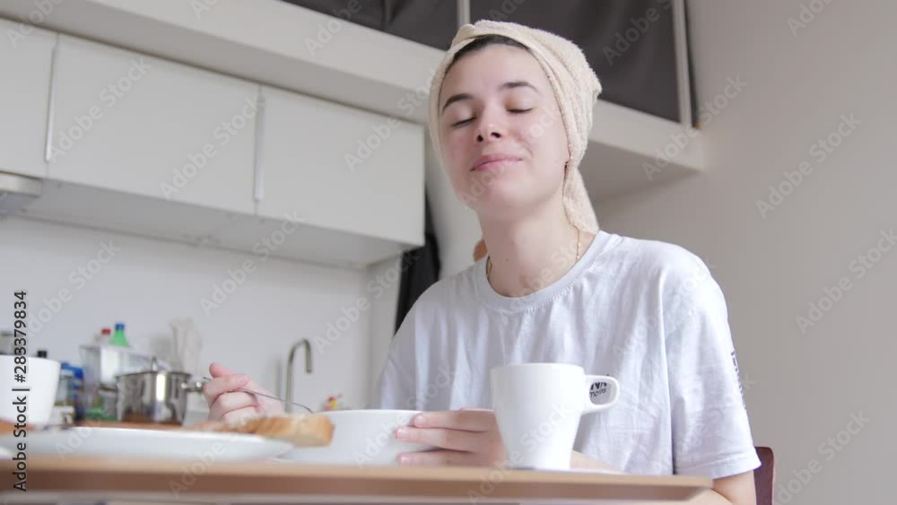 A young girl with a towel on her head is having breakfast, eating morning cereal, after a shower, looking out the window, smiling. White T-shirt, minimalism, stylish, no makeup A young girl with a towel on her head is having breakfast, eating morning cereal, after a shower, looking out the window, smiling. White T-shirt, minimalism, stylish, no makeup