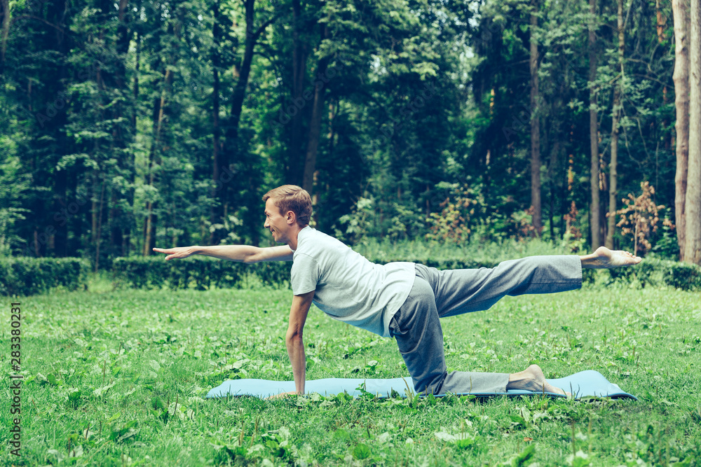 Young attractive man in a gray T-shirt and sweatpants doing yoga pose in the park. Keeps balance on one leg Young attractive man in a gray T-shirt and sweatpants doing yoga pose in the park. Keeps balance on one leg