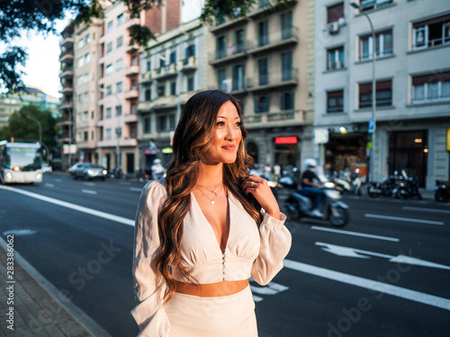 Summer sunny lifestyle portrait of young stylish asian girl on the street of big modern city. She wears white dress. women business