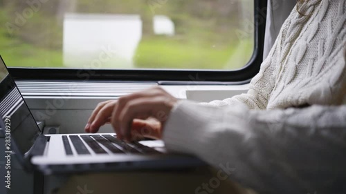 Young girl travelling on train in daytime. Woman sitting near window, working on computer. Detail view female hands opening notebook and starts typing on keyboard. Concept freelancer job in journey 