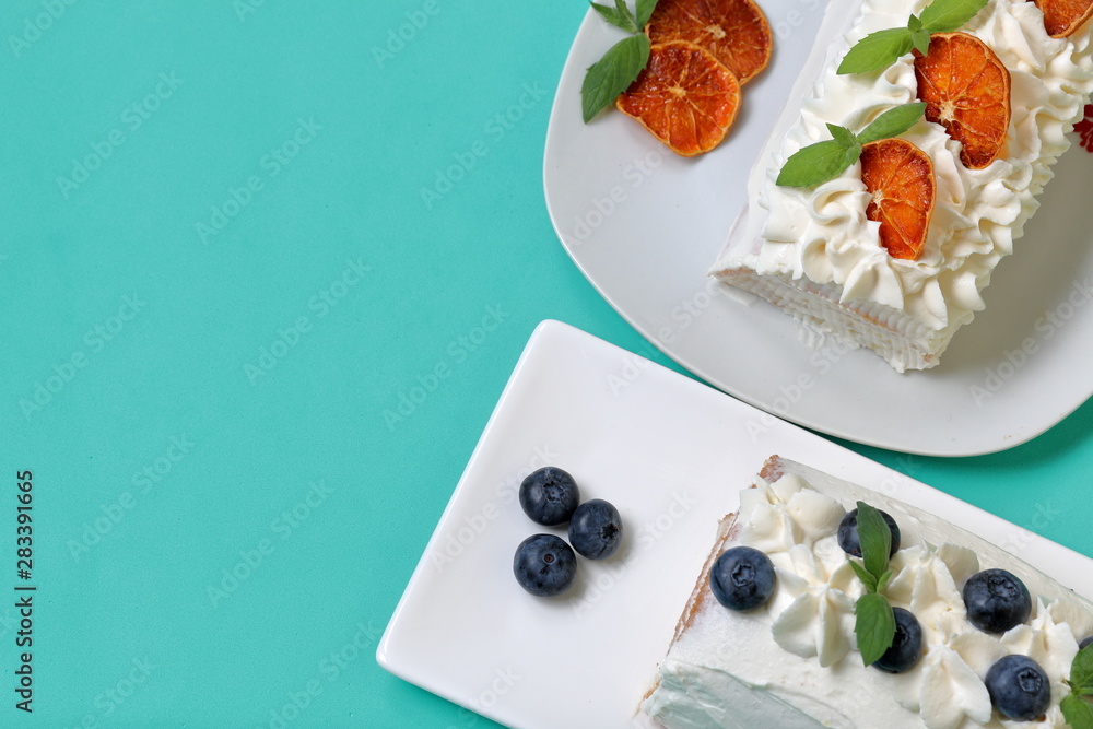 Sponge cake coated with cream and garnished with dried blueberries and fresh mint leaves. Also dry slices of orange. Against the background of mint color. Sponge cake coated with cream and garnished with dried blueberries and fresh mint leaves. Also dry slices of orange. Against the background of mint color.