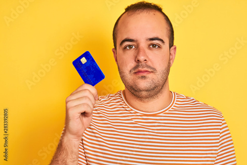 Young man holding blue credit card standing over isolated yellow background with a confident expression on smart face thinking serious