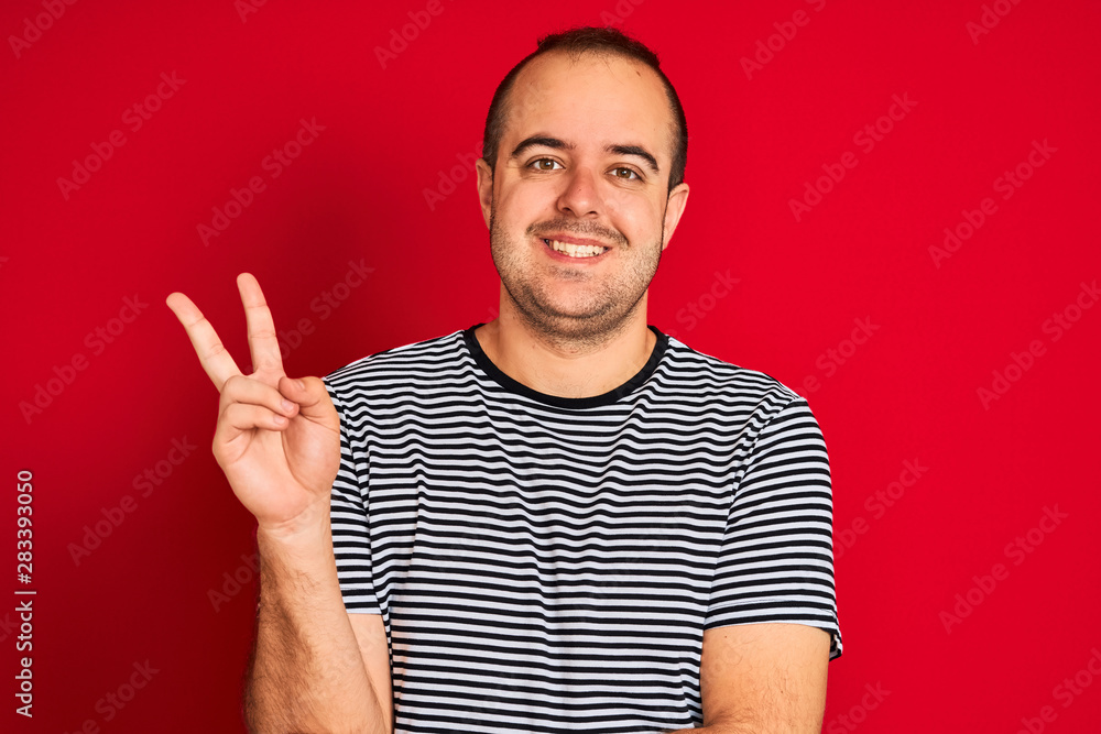 Fototapeta premium Young man wearing striped navy t-shirt standing over isolated red background smiling with happy face winking at the camera doing victory sign. Number two.