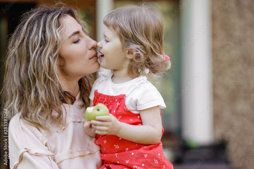 Young mother holds her little daughter on her hands and kisses her in the garden on a warm summer day Young mother holds her little daughter on her hands and kisses her in the garden on a warm summer day