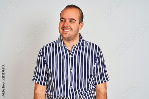 Young man wearing blue striped shirt standing over isolated white background looking away to side with smile on face, natural expression. Laughing confident.