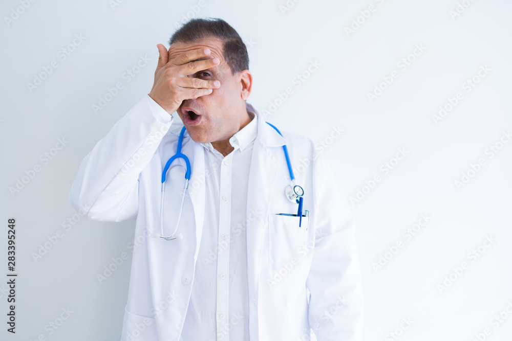 Middle age doctor man wearing stethoscope and medical coat over white background peeking in shock covering face and eyes with hand, looking through fingers with embarrassed expression. Middle age doctor man wearing stethoscope and medical coat over white background peeking in shock covering face and eyes with hand, looking through fingers with embarrassed expression.