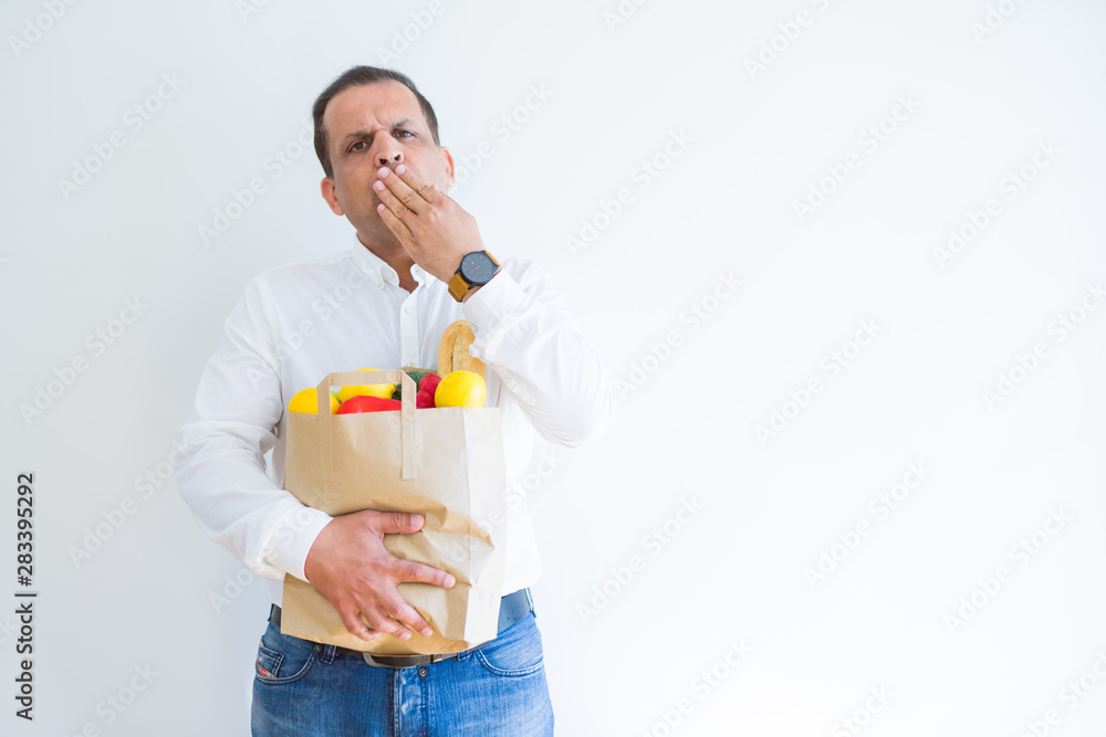 Middle age man holding groceries shopping bag over white background cover mouth with hand shocked with shame for mistake, expression of fear, scared in silence, secret concept Middle age man holding groceries shopping bag over white background cover mouth with hand shocked with shame for mistake, expression of fear, scared in silence, secret concept