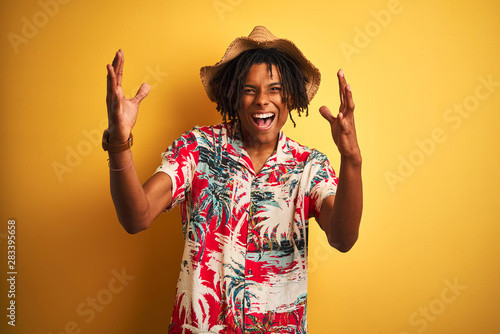 Afro american man with dreadlocks wearing floral shirt and hat over isolated yellow background celebrating crazy and amazed for success with arms raised and open eyes screaming excited. Winner concept