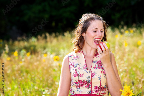 woman in dirndl standing in field of flowers and eating an apple