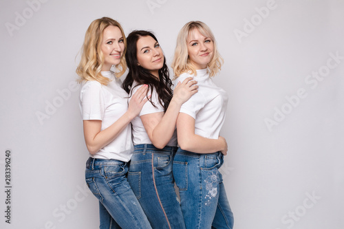 Side view of three beautiful young women standing one by one. Blonde and brunette girls in white t-shirts and jeans posing at camera. Happy friends in casual clothes smiling being close to each other.
