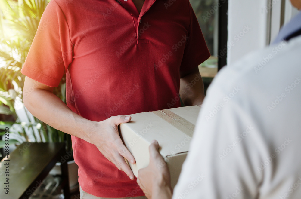 Obraz premium hand of young delivery service man in red uniform with man customer receiving parcel post box from courier at home, cargo shipping, express delivery service, online shopping and logistics concept