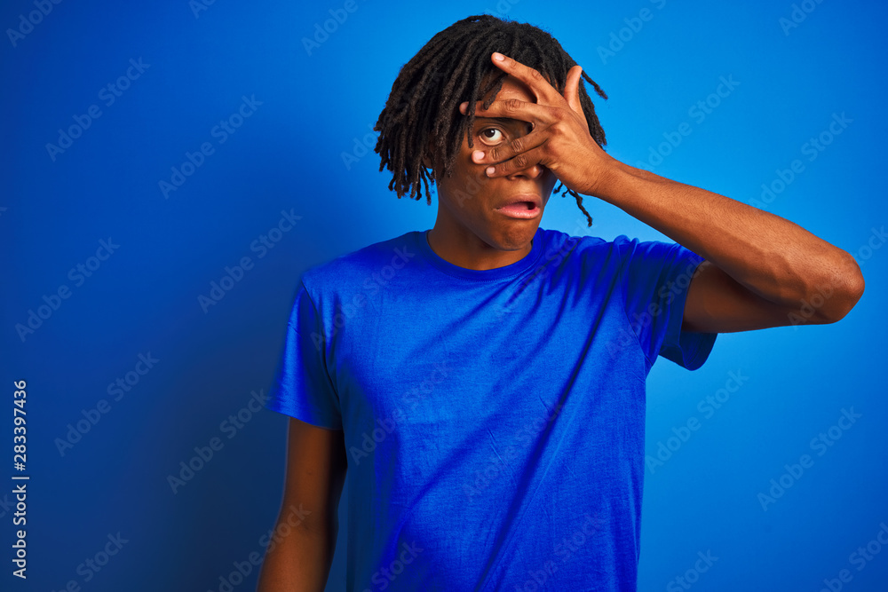 Afro american man with dreadlocks wearing t-shirt standing over isolated blue background peeking in shock covering face and eyes with hand, looking through fingers with embarrassed expression. Afro american man with dreadlocks wearing t-shirt standing over isolated blue background peeking in shock covering face and eyes with hand, looking through fingers with embarrassed expression.