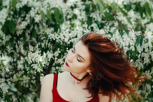 Fototapeta Naklejka Na Ścianę i Meble -  Beautiful young woman enjoying smell in a flowering spring garden.Beutiful tree , flowers. Red hair, blue eyes and red dress,holds the flowers in their hands.