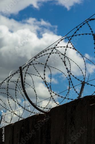 Strained sharp barbed wire on top of a fence silhouette. Blue sky with white clouds. Border of the forbidden territory. Restricted area. Wavy wire with prickles.