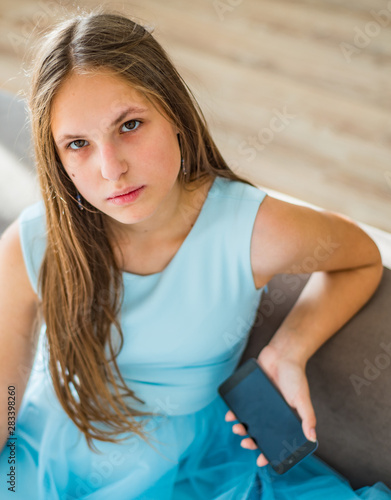 portrait of young teenager brunette girl with long hair sitting on sofa at home in blue dress with smartphone