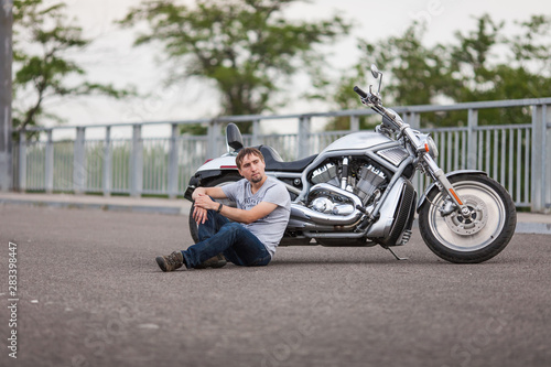 Handsome rider man sitting near classic style cruiser motorbike on an open road. Brutal young man sitting at powerful shiny style cruiser motorbike on highway