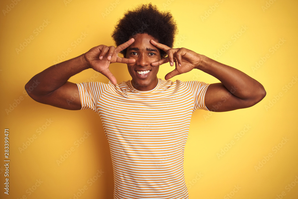 American man with afro hair wearing striped t-shirt standing over isolated yellow background Doing peace symbol with fingers over face, smiling cheerful showing victory American man with afro hair wearing striped t-shirt standing over isolated yellow background Doing peace symbol with fingers over face, smiling cheerful showing victory