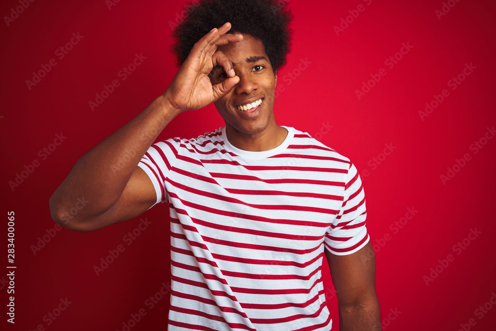 Young african american man with afro hair wearing striped t-shirt over isolated red background doing ok gesture with hand smiling, eye looking through fingers with happy face. Young african american man with afro hair wearing striped t-shirt over isolated red background doing ok gesture with hand smiling, eye looking through fingers with happy face.