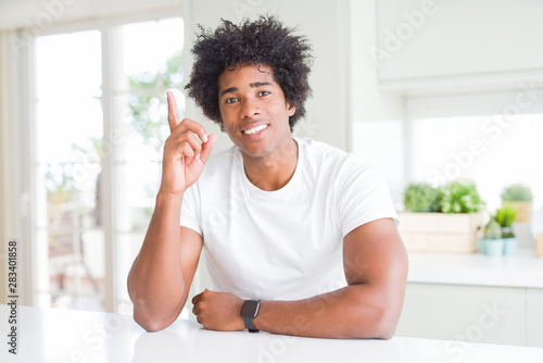 Young african american man wearing casual white t-shirt sitting at home showing and pointing up with finger number one while smiling confident and happy.