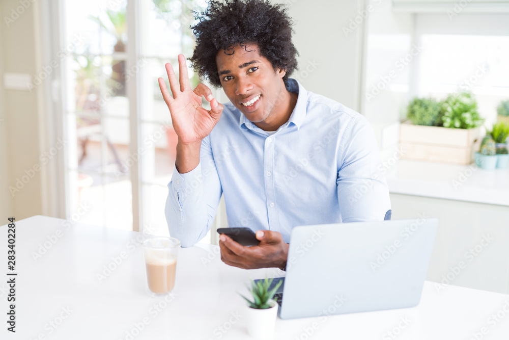 African American business man using smartphone and laptop doing ok sign with fingers, excellent symbol African American business man using smartphone and laptop doing ok sign with fingers, excellent symbol